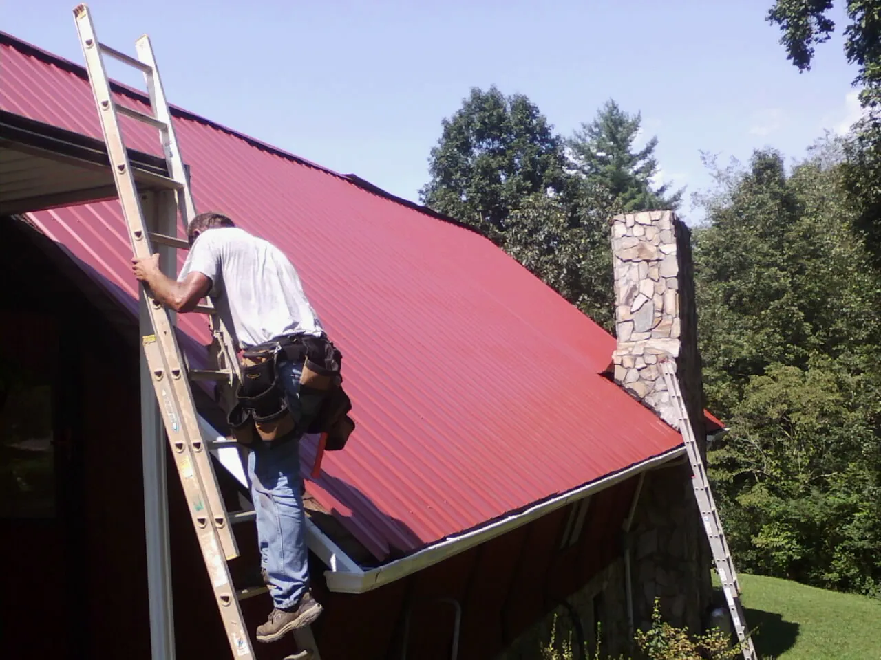 Roofer on a ladder beside a red metal roof and stone chimney — Soffit & Fascia Repair by Roofing Brothers in Poseyville